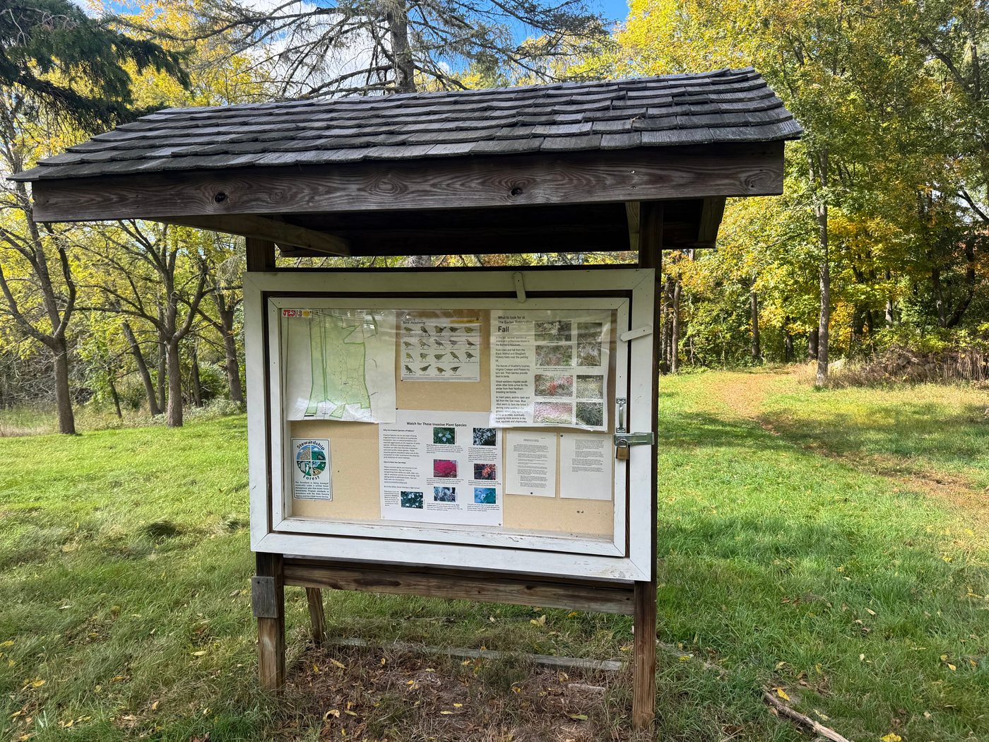 The shingle-roofed trailhead kiosk at Barber Reservation, with a trail map and seasonal nature notes, on a mowed field edged with fall trees