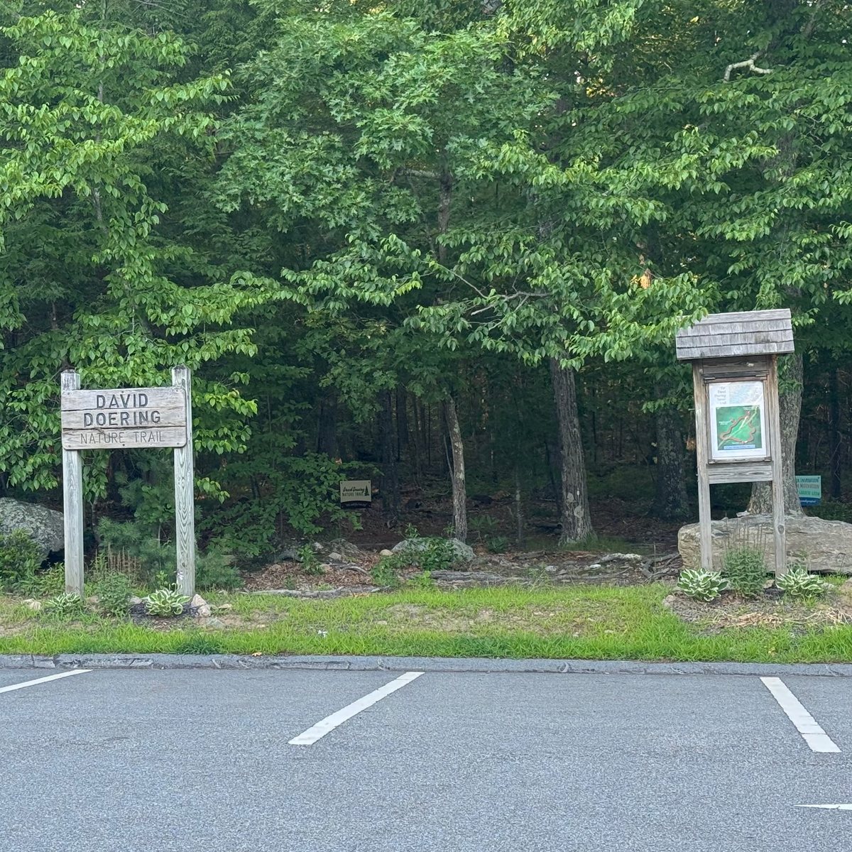 The wooden David Doering Nature Trail sign and a shingle-roofed trail map kiosk at the edge of the woods, beside a small parking lot