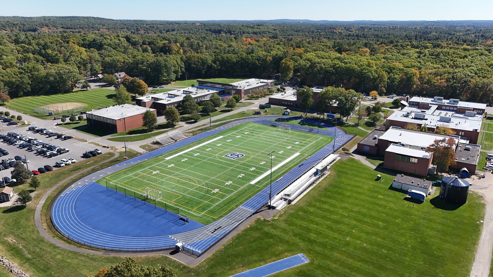 Aerial view of the Dover-Sherborn High School and Middle School campus, with the blue track and turf field, the school buildings, and woods beyond