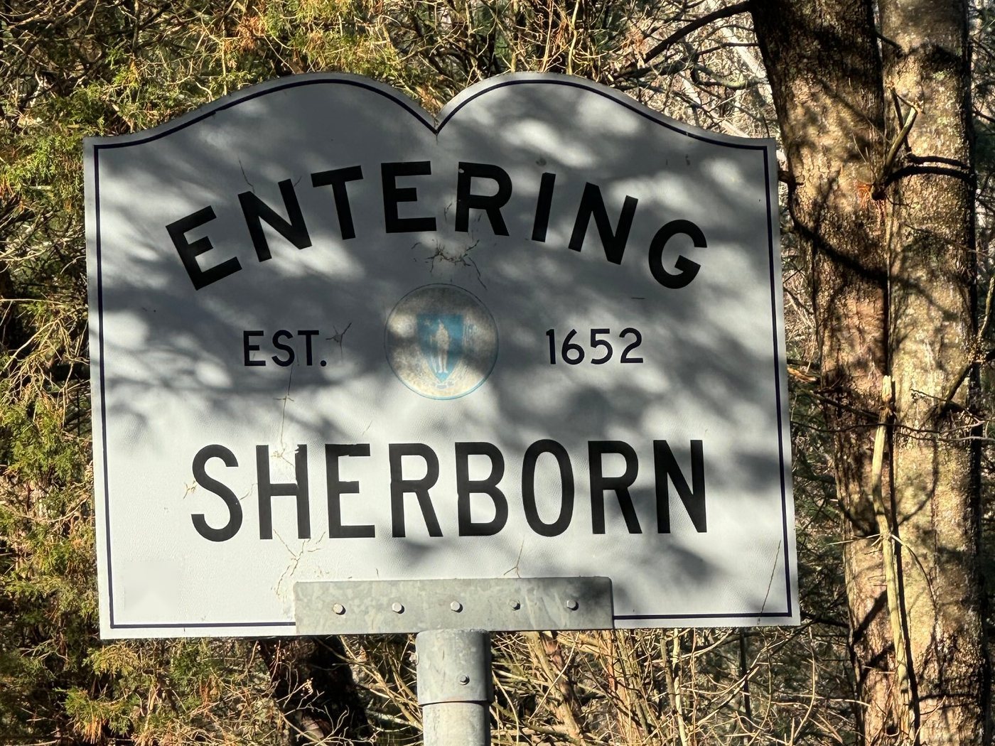 A white Entering Sherborn town line sign reading Est. 1652, with the Massachusetts seal, in dappled sunlight against bare trees