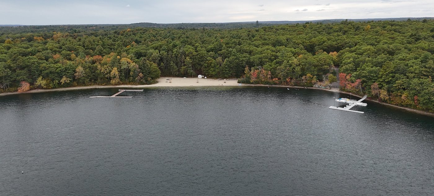 Aerial view of the sandy Farm Pond town beach and swim dock, with the Sherborn Yacht Club docks to the right and fall woods all around