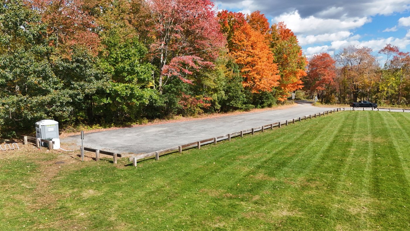 The gravel parking lot at Fessenden Fields, lined with a low wooden fence, with a portable toilet at one end