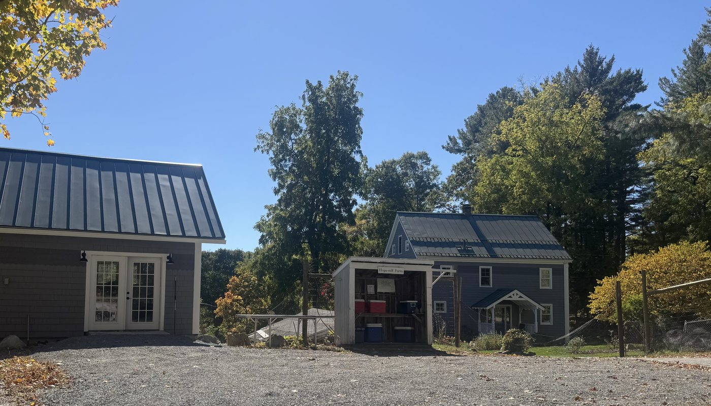 The Hopestill Farm stand, a small open shed with coolers, between a barn and a blue farmhouse