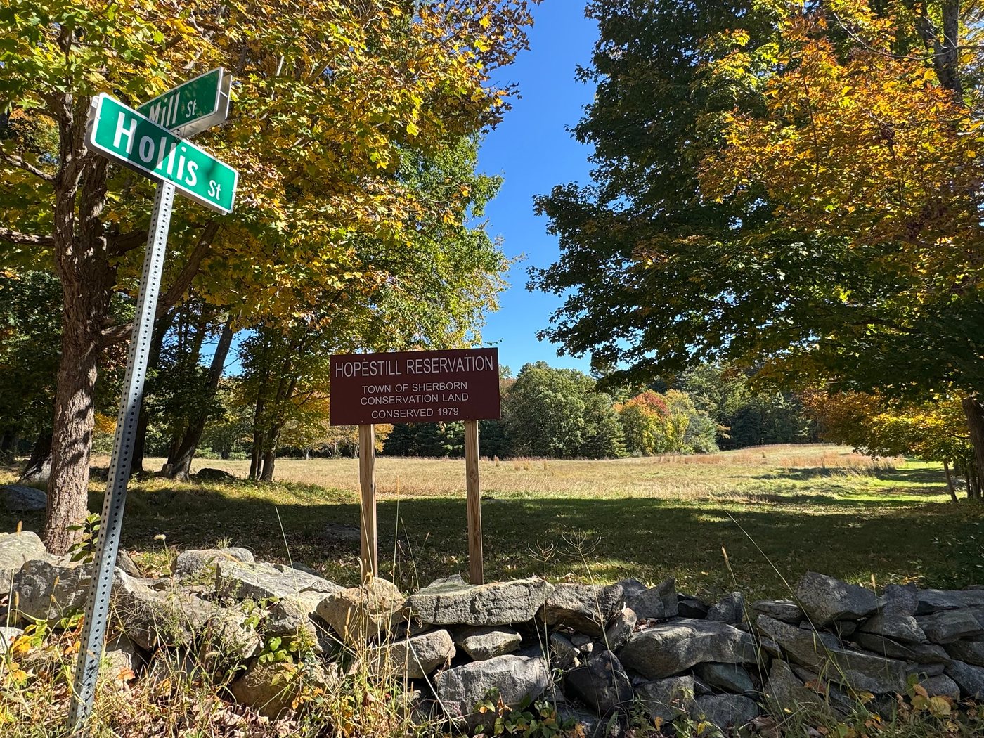 The Hopestill Reservation sign at the corner of Hollis Street and Mill Street, behind an old stone wall, with an open meadow and fall maples beyond