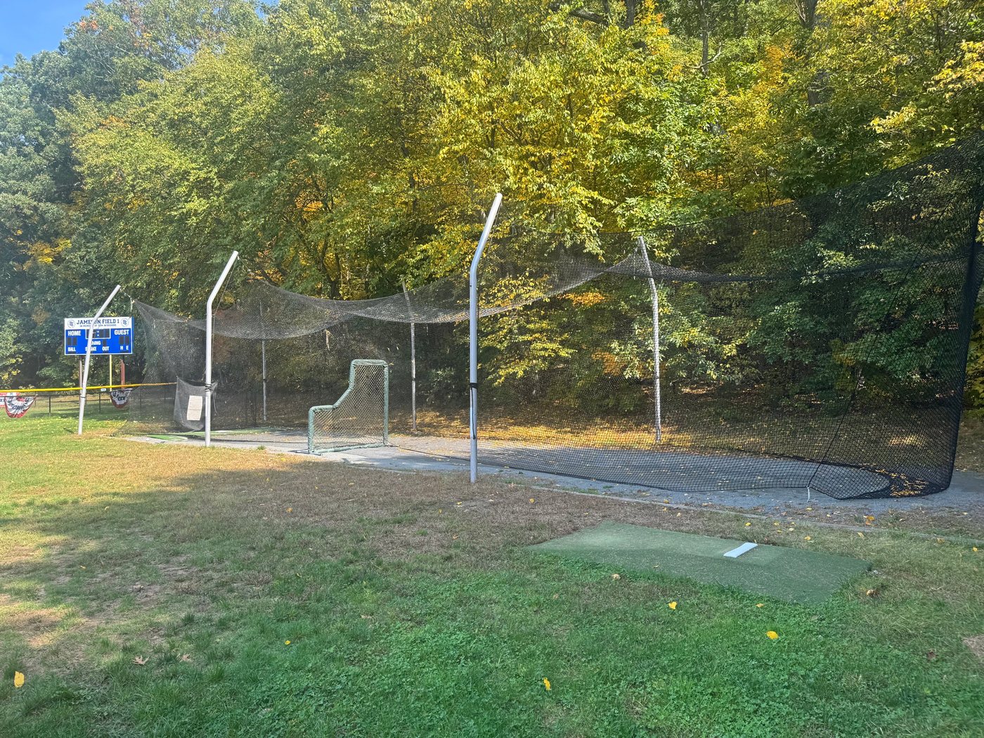 The netted batting cages at Jameson Fields under fall trees, with a pitching mat in front and the Field 1 scoreboard at left