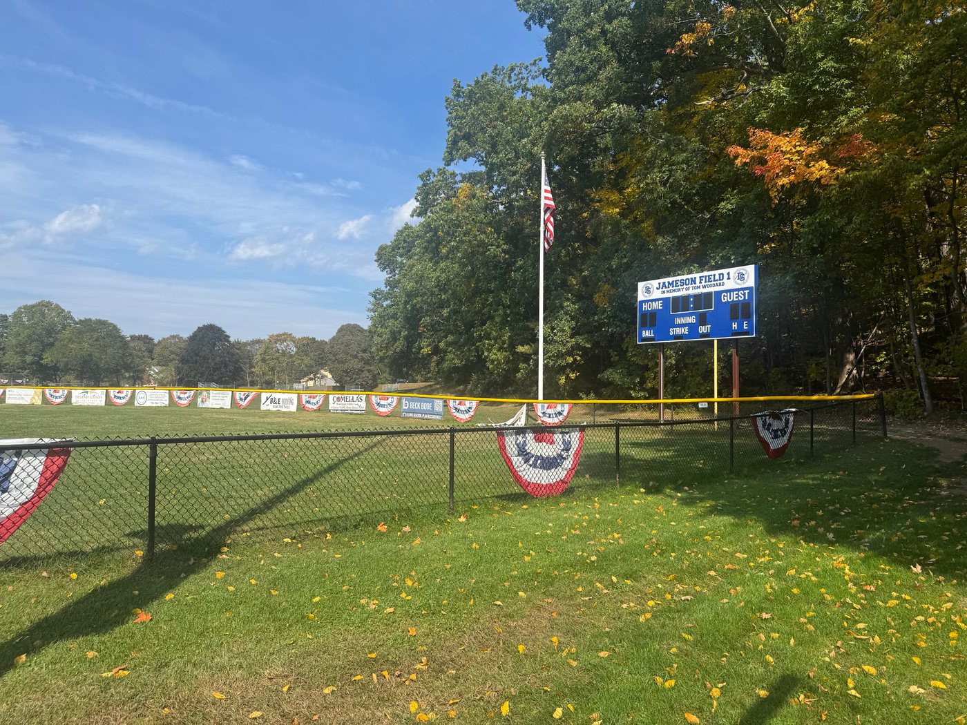 The blue Jameson Field 1 scoreboard, in memory of Tom Woodard, beside the flagpole and the bannered outfield fence