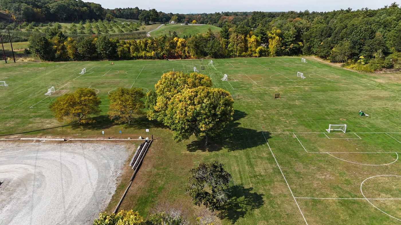 Laurel Farm Fields from above, with the gravel parking lot, shade trees, and lined soccer fields, and Dowse Orchards on the hill beyond
