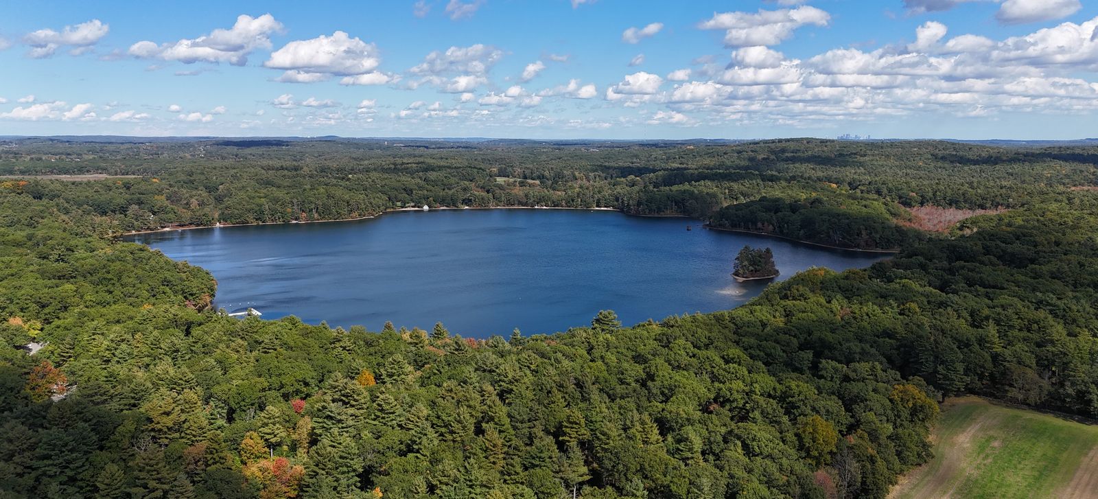 Aerial view of Farm Pond surrounded by woods, with an open field in the foreground and the Boston skyline on the far horizon
