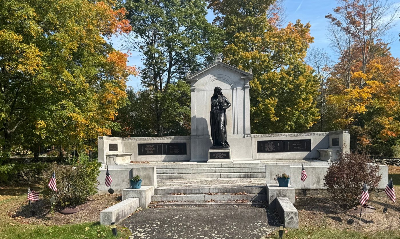 Sherborn’s war memorial in fall, with Cyrus Dallin’s bronze Memory statue at the center of a granite monument lined with bronze name plaques and small American flags