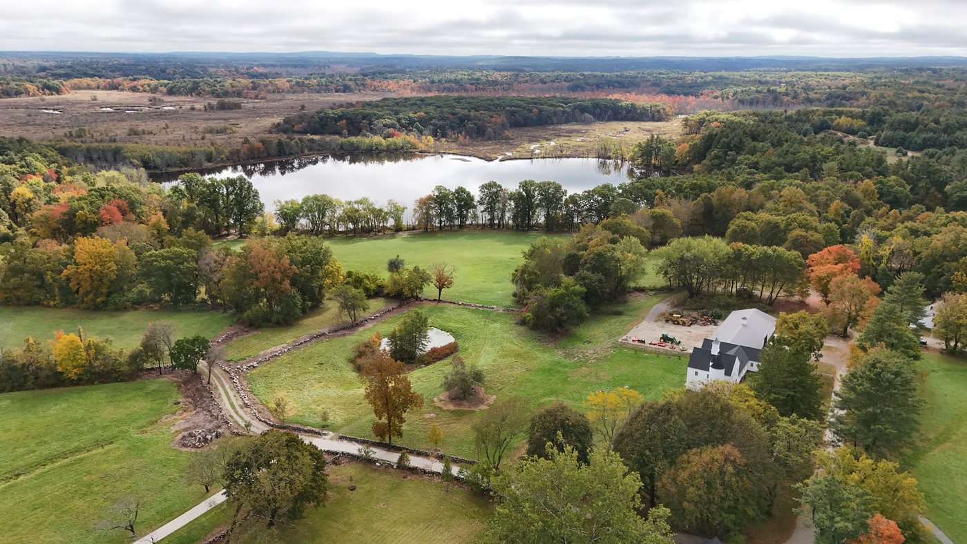 Millborn Farm from above in fall, with the white barn, a small pond, stone-walled hay fields, and the Charles River and its meadows beyond