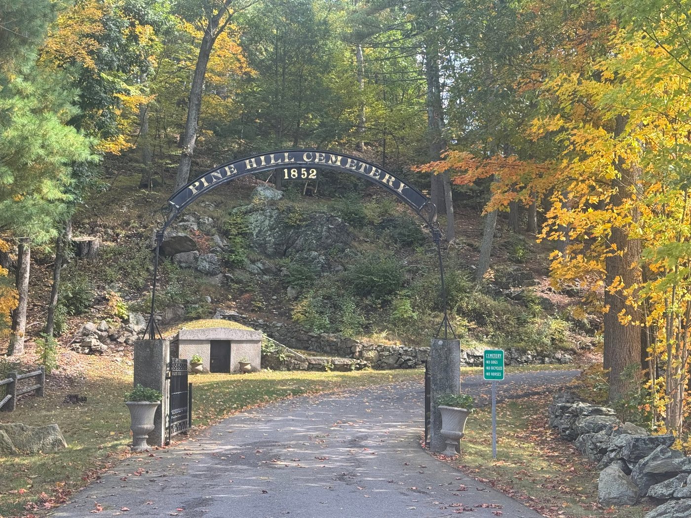 The iron Pine Hill Cemetery arch, dated 1852, on granite posts with urns, framed by fall maples, with a granite tomb in the hillside beyond