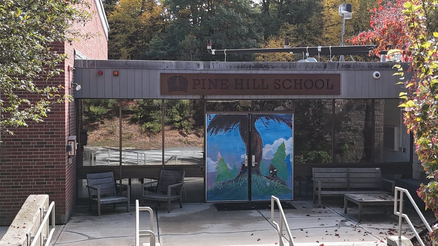 The front entrance of Pine Hill School, with a painted mural of a tall tree on the doors beneath the school’s name, benches on the patio, and fall leaves