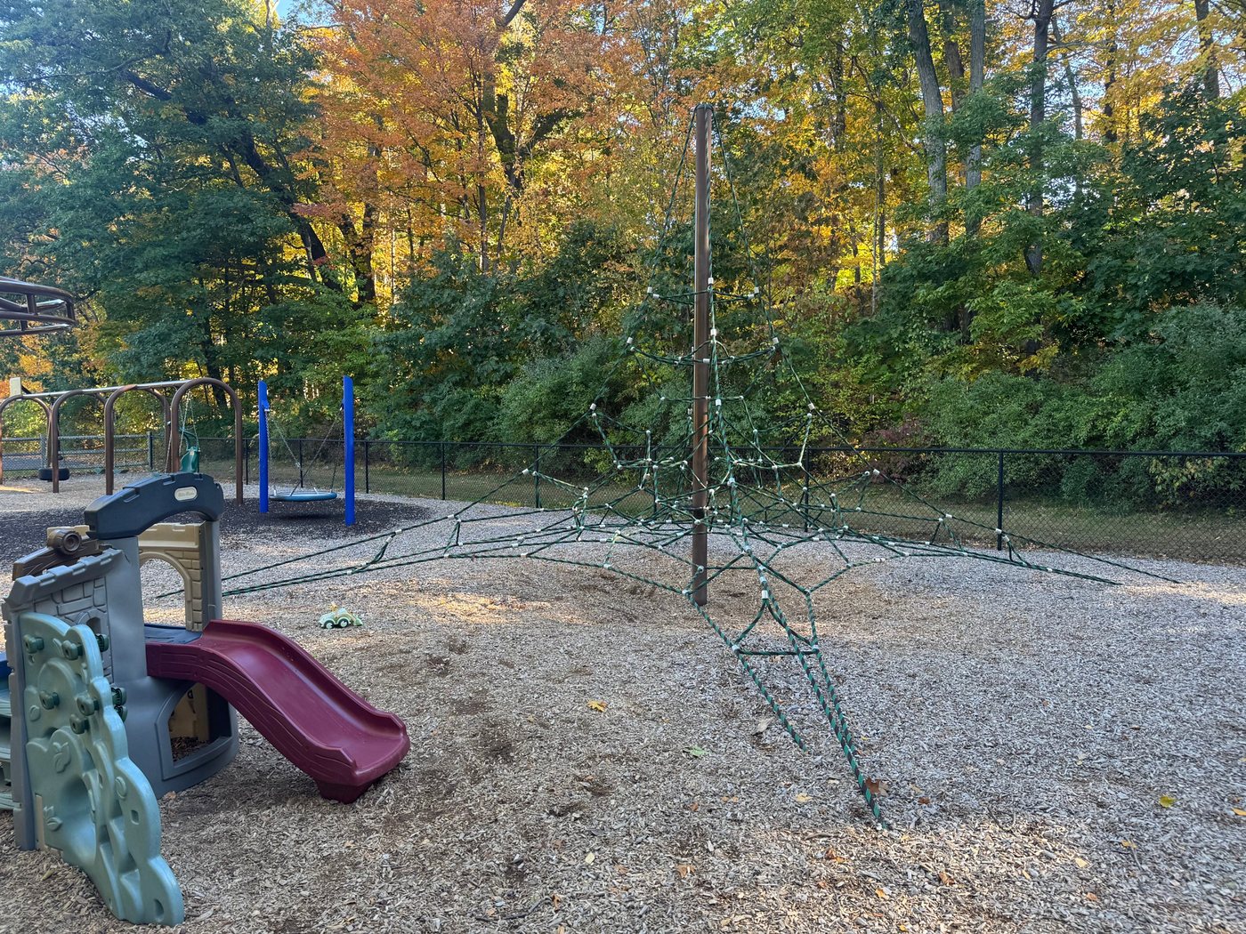 A tall rope climbing net at Sherborn Playground, with a saucer swing and a small slide nearby under fall foliage