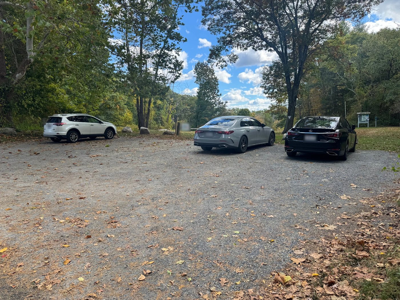 The gravel parking lot off Forest Street at Rocky Narrows, edged with trees, with a trail kiosk on the right