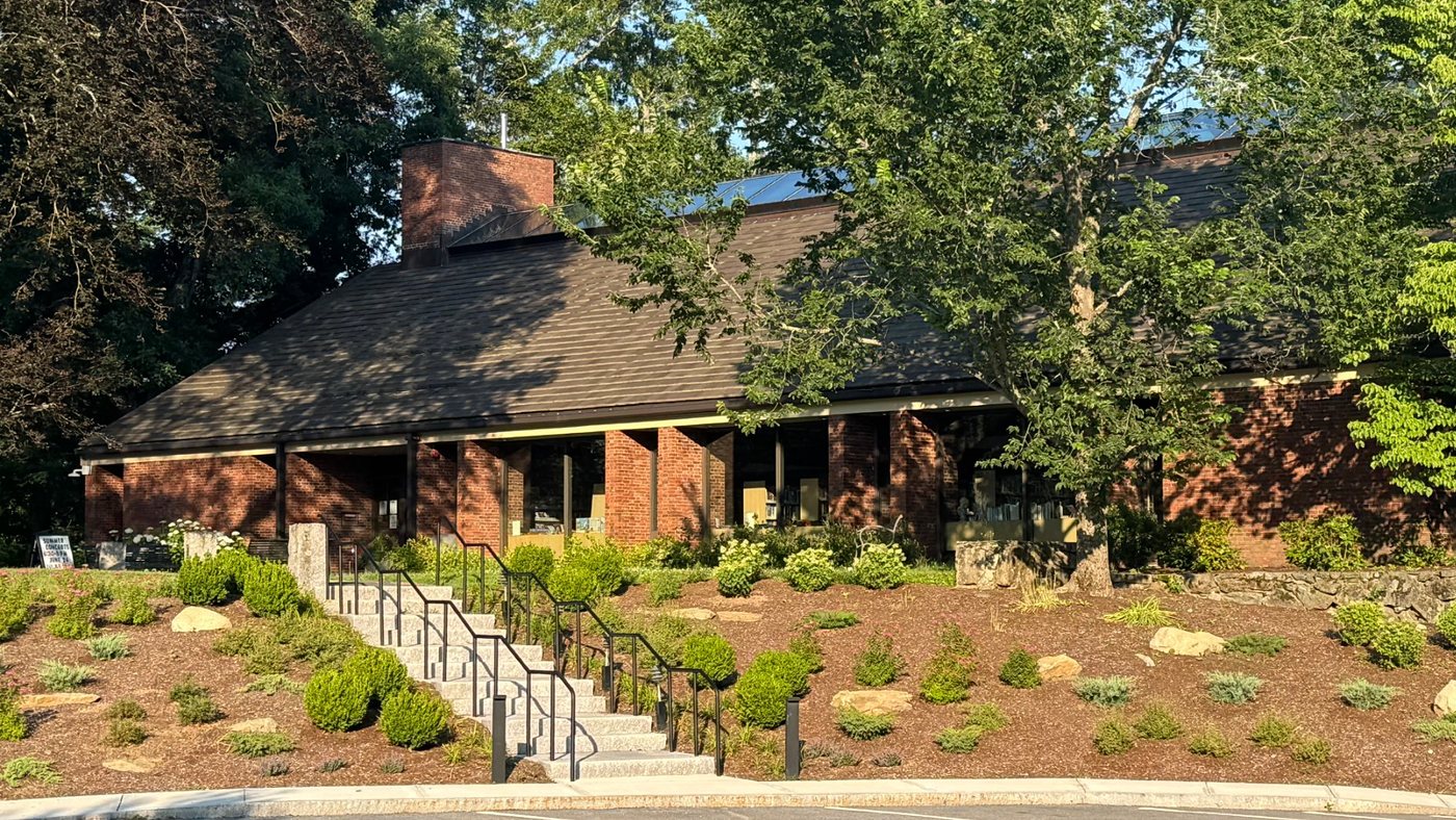 The brick Sherborn Library with its long sloped roof, landscaped front slope, and granite steps, under summer trees