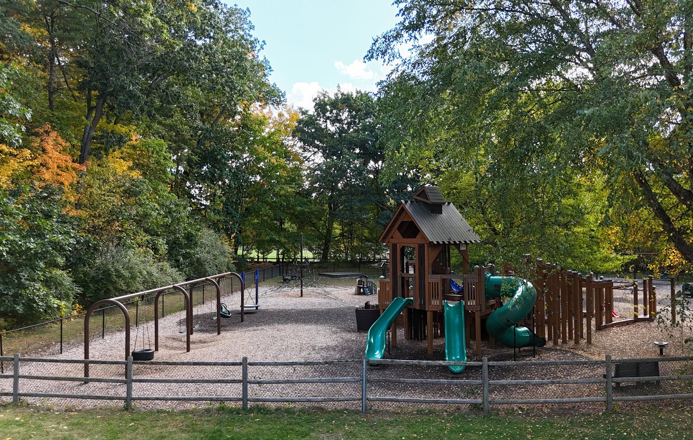 Sherborn Playground, with swings, a rope climber, and a wooden play structure with green slides, behind a split-rail fence under fall trees