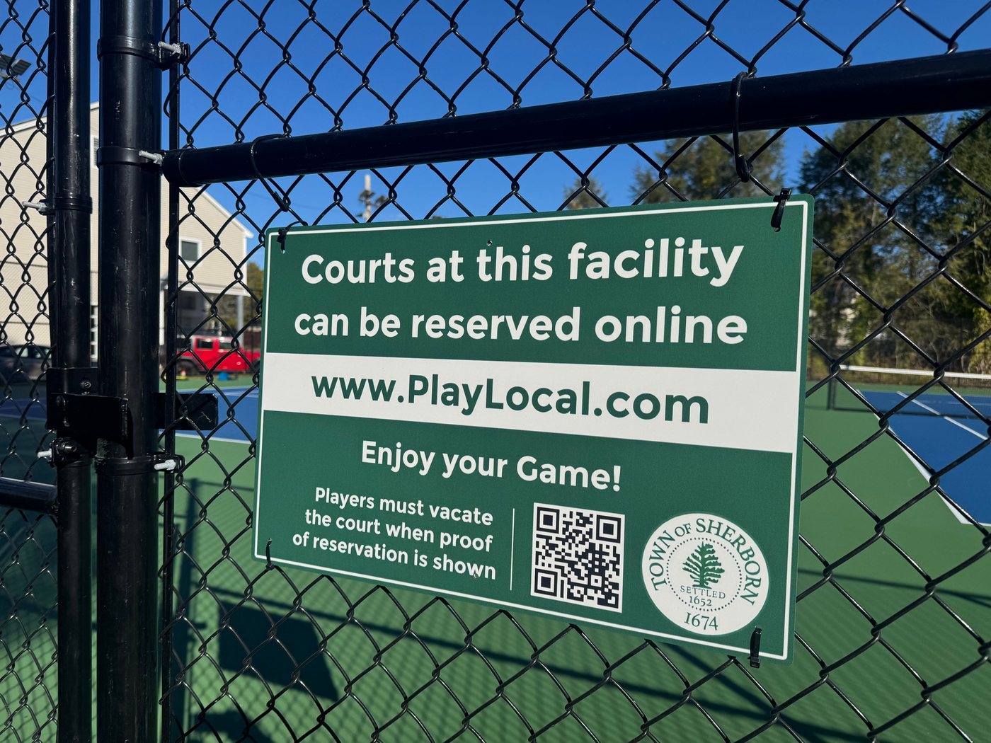 The green Town of Sherborn sign on the court fence: courts at this facility can be reserved online at PlayLocal.com