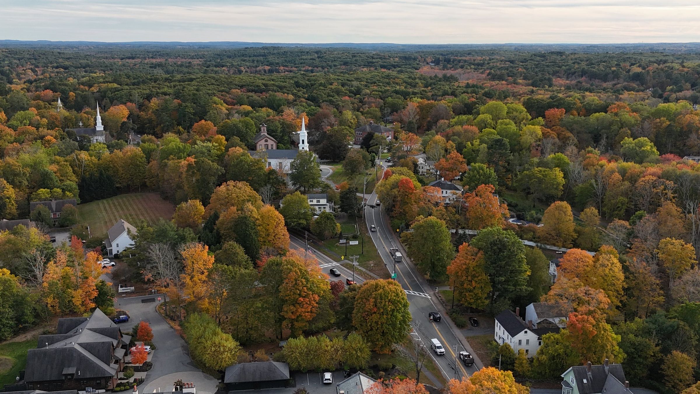 Aerial view of the center of Sherborn in fall, with two white spires rising above trees in orange, red, and gold