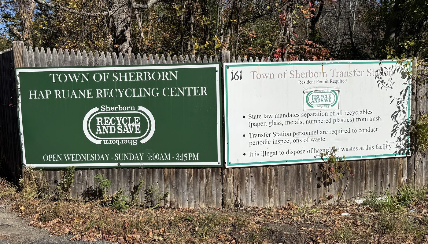 Entrance signs for the Town of Sherborn Hap Ruane Recycling Center and Transfer Station, on a wooden fence under fall foliage